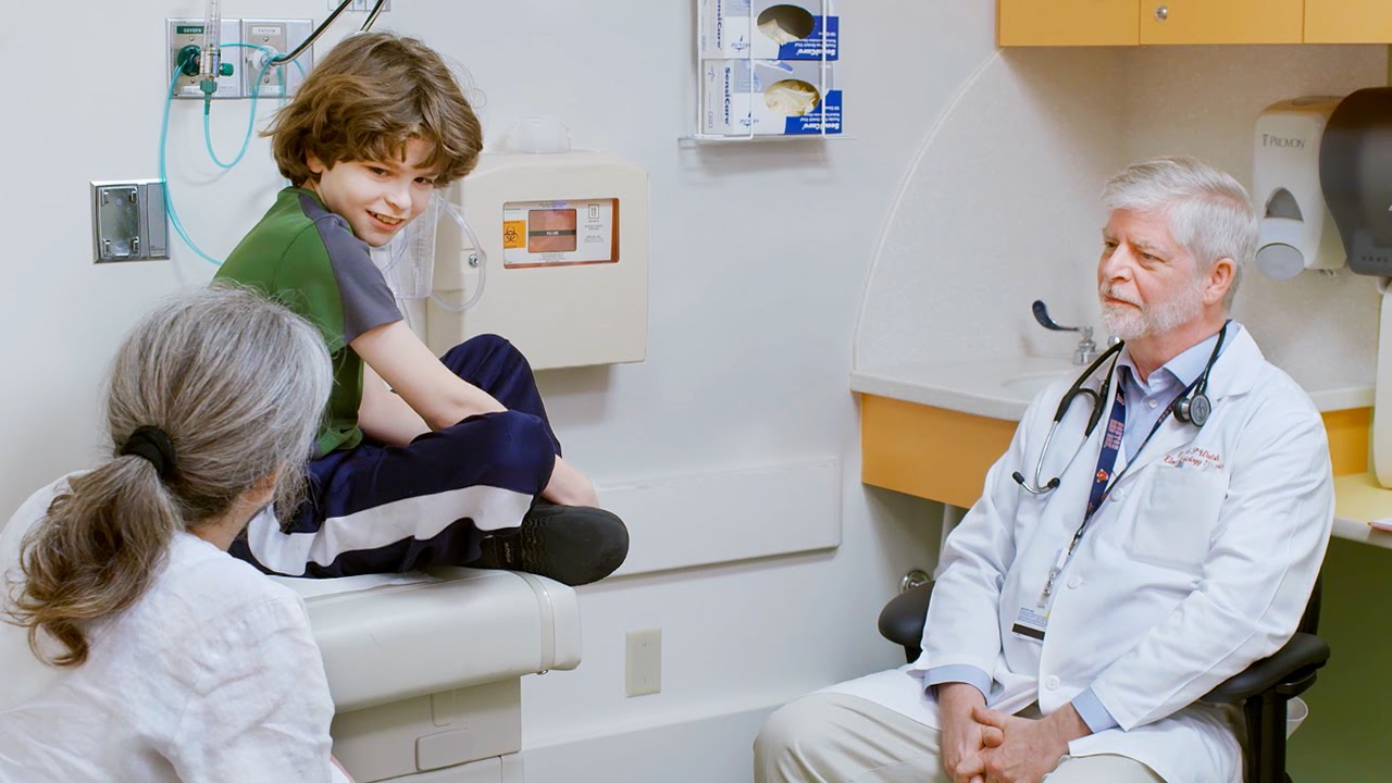 Boy sitting on exam room table (center) speaks with female clinician with hair in a pony tail (left) and male clinician with a beard (right)