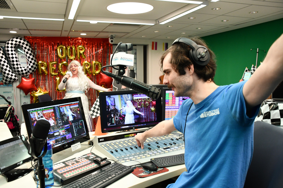 Man behind computer holds up arms as woman performs live on camera