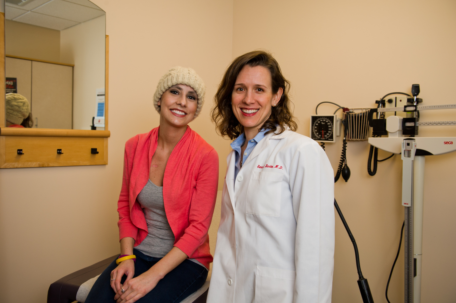 Woman wearing hat sits on exam table next to doctor, standing