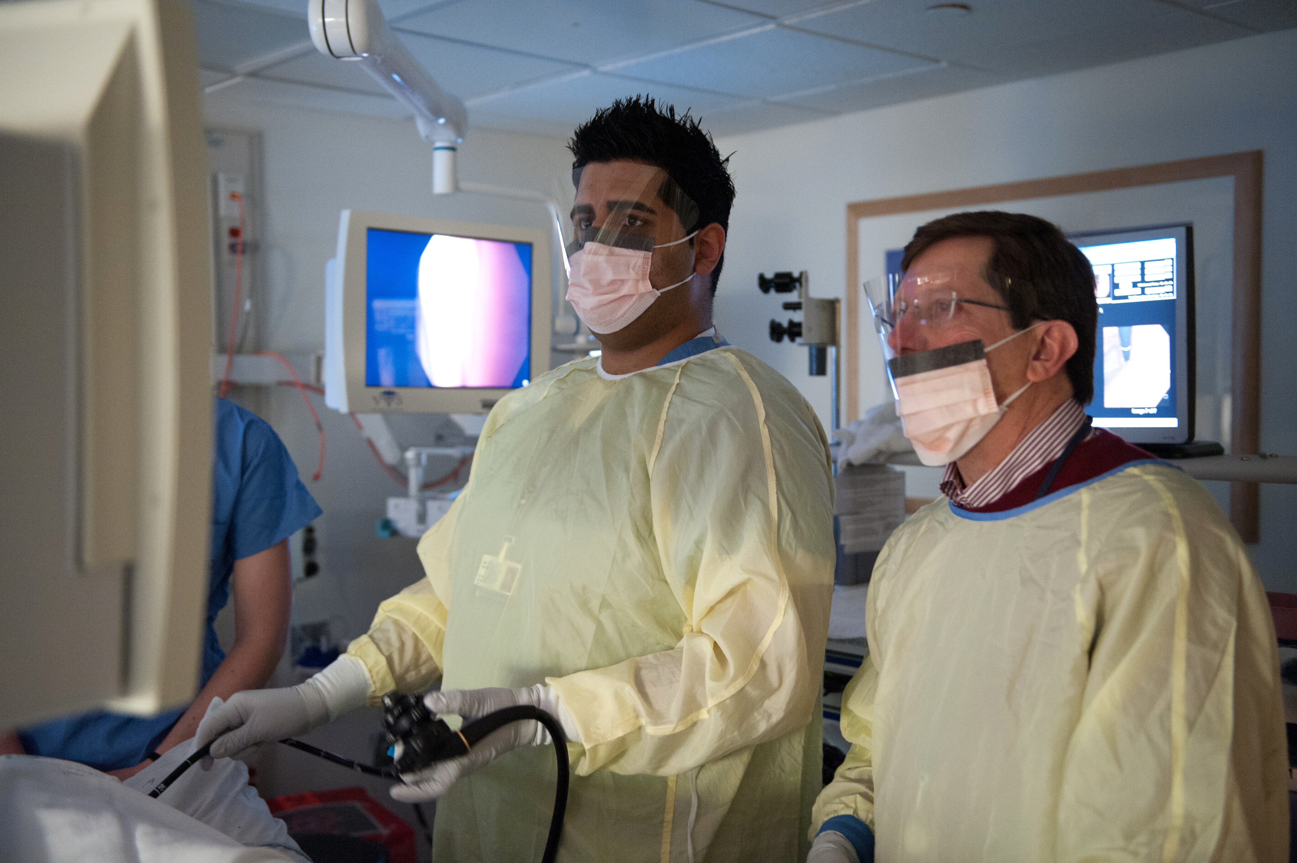 Two male clinicians in yellow gowns, face masks, and eye shields look toward a monitor