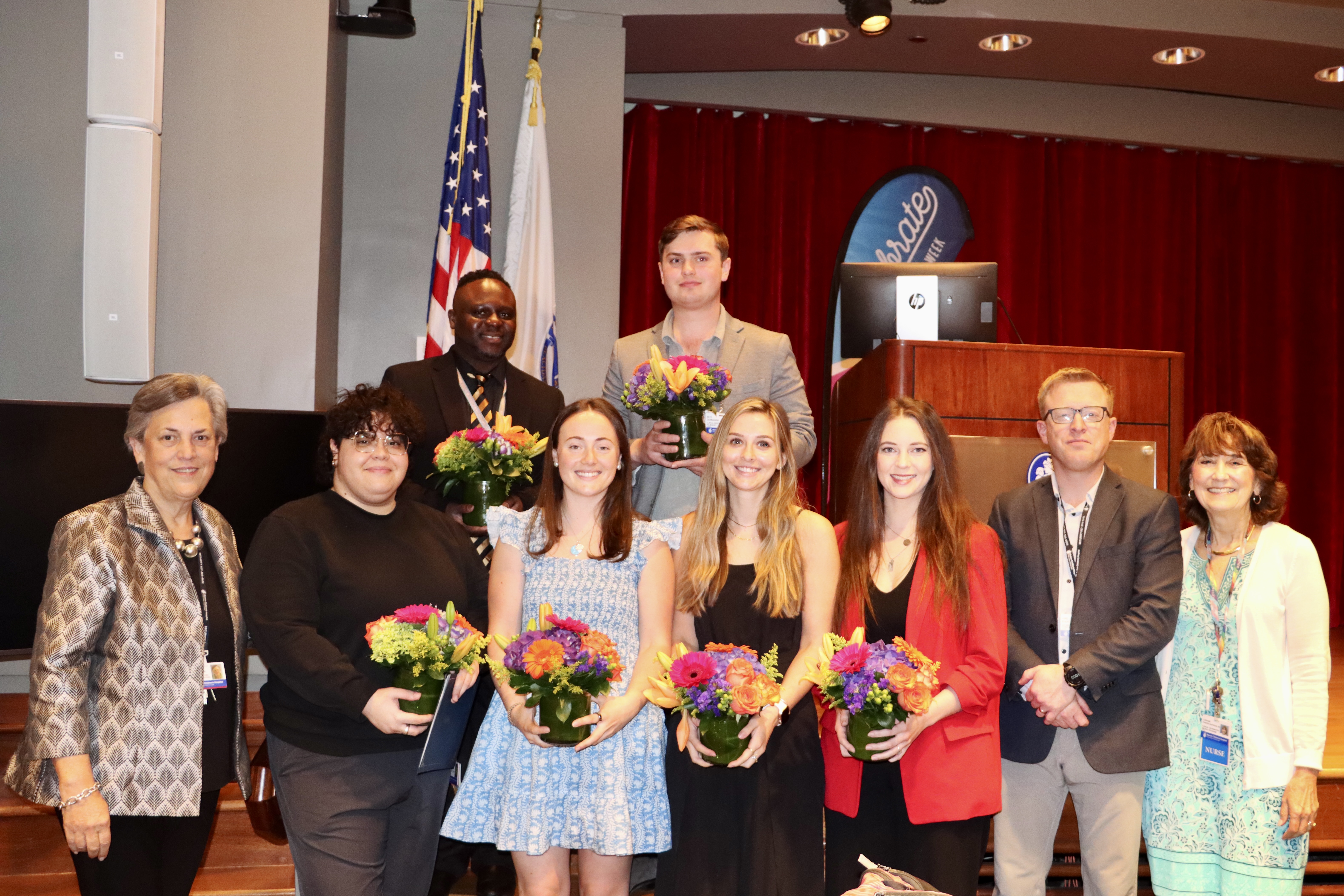 A group of clinical team assistant exemplars stand on a stage, posing with flowers.