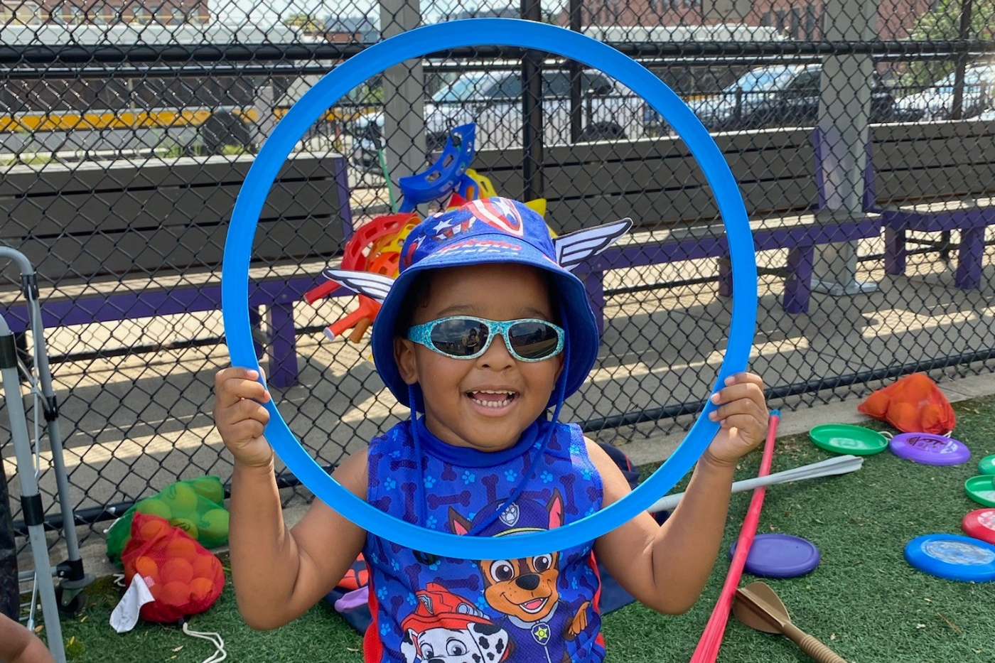 Child wearing sunglasses and helmet holds up a hula hoop.