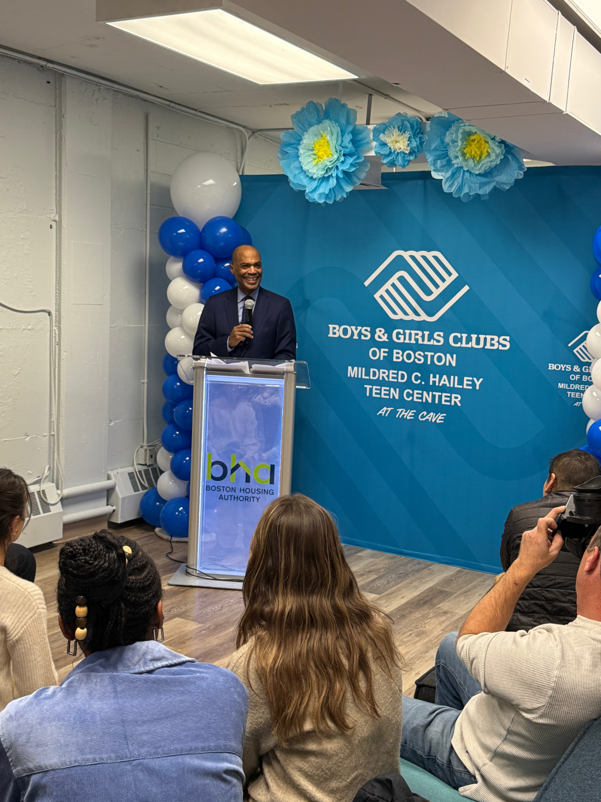 Tall man speaks behind podium. Sign for Boys & Girls Club of Boston in background. Small audience watches.