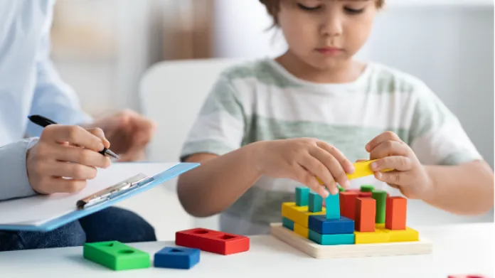A young child plays with colorful wooden blocks while an adult, holding a clipboard and pen, observes and takes notes.