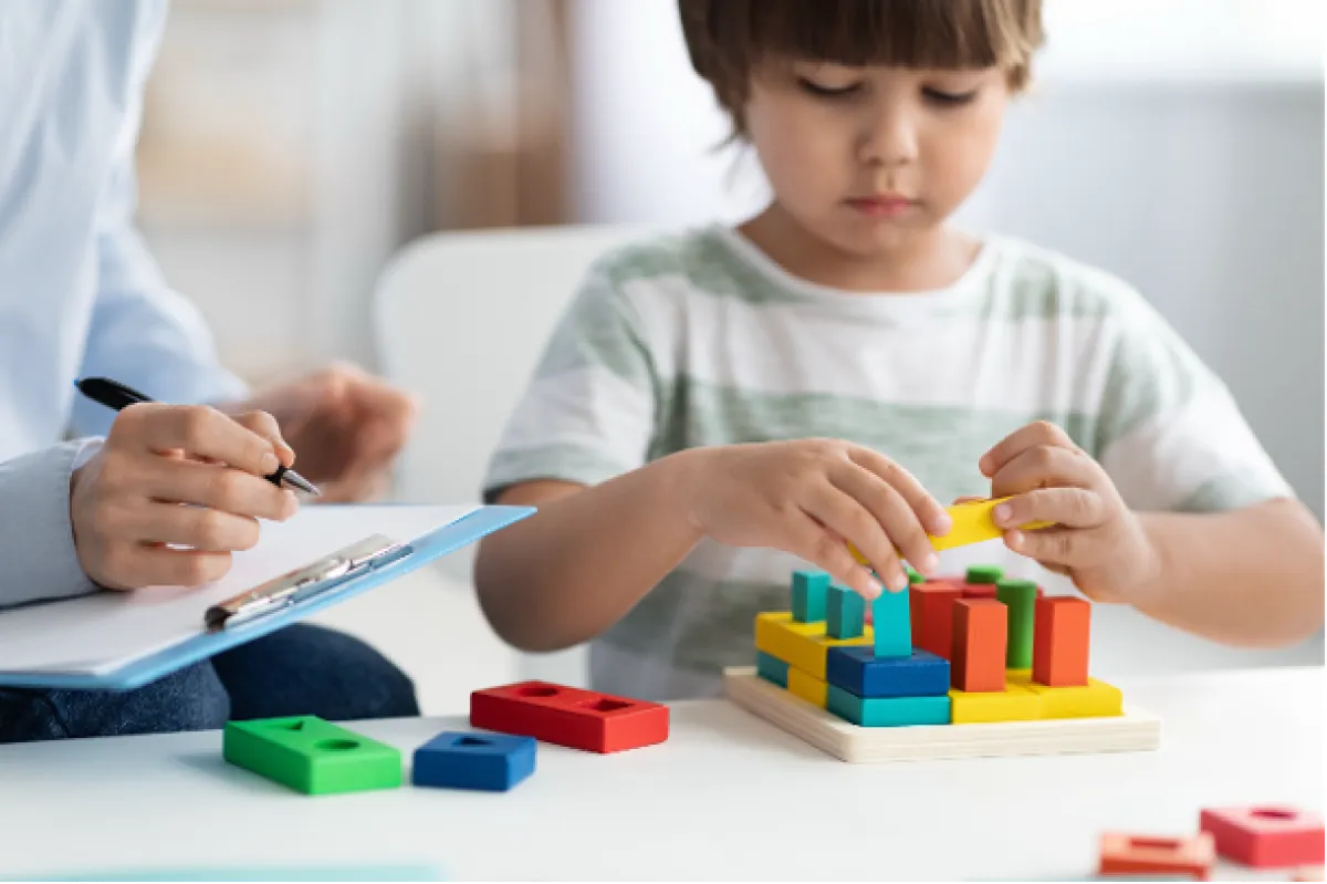 A young child plays with colorful wooden blocks while an adult, holding a clipboard and pen, observes and takes notes.