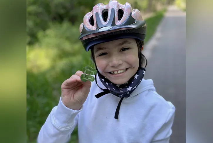 Girl on bicycle wearing bicycle helmet smiles, holds a four-leaf clover