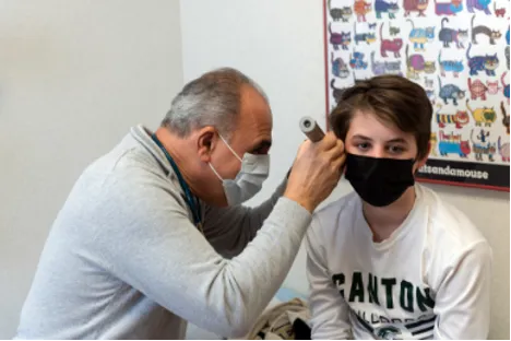 A doctor in a mask shines a light into a masked patient's ear.
