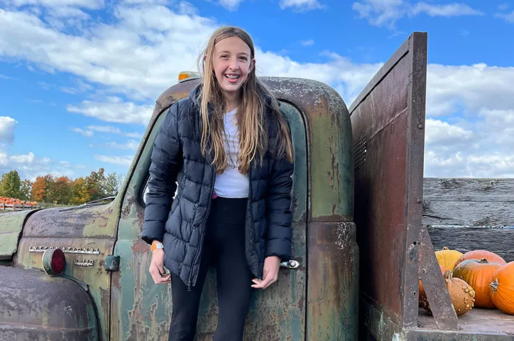 Preteen blonde-haired girl stands in front of tractor that also carries pumpkins
