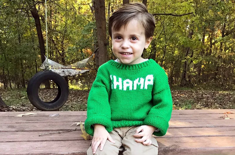 Little boy wearing a sweater while sitting on a log smiles for the camera.