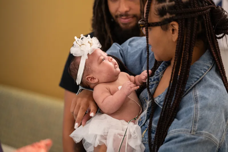 Woman holds young child wearing bow on head. Man in background looks on.