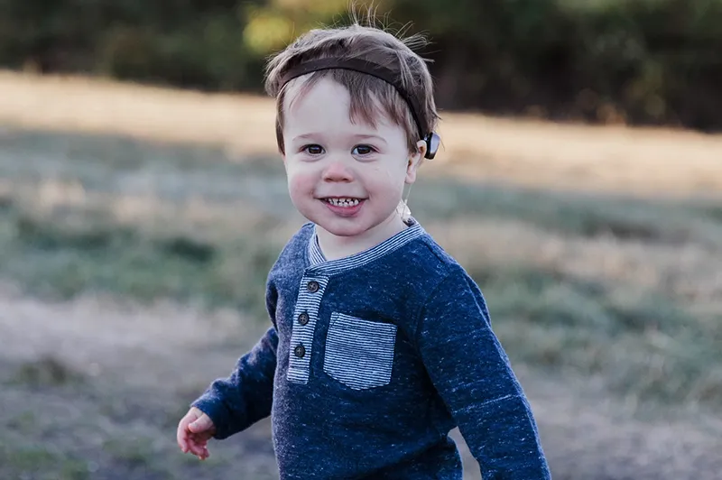 Small boy wearing blue long-sleeve shirt and bone-anchored hearing system smiles for photo