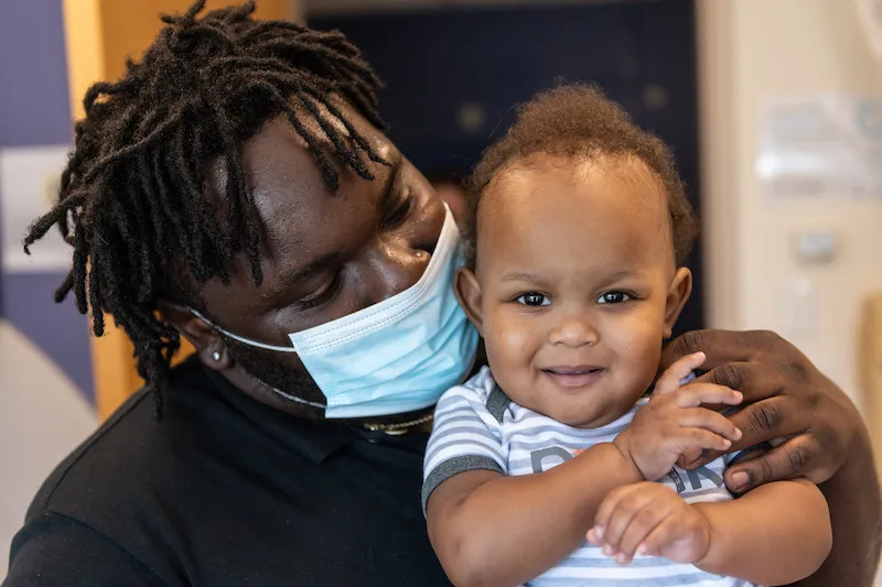 Man with braided hair and wearing mask holds young boy in his arms
