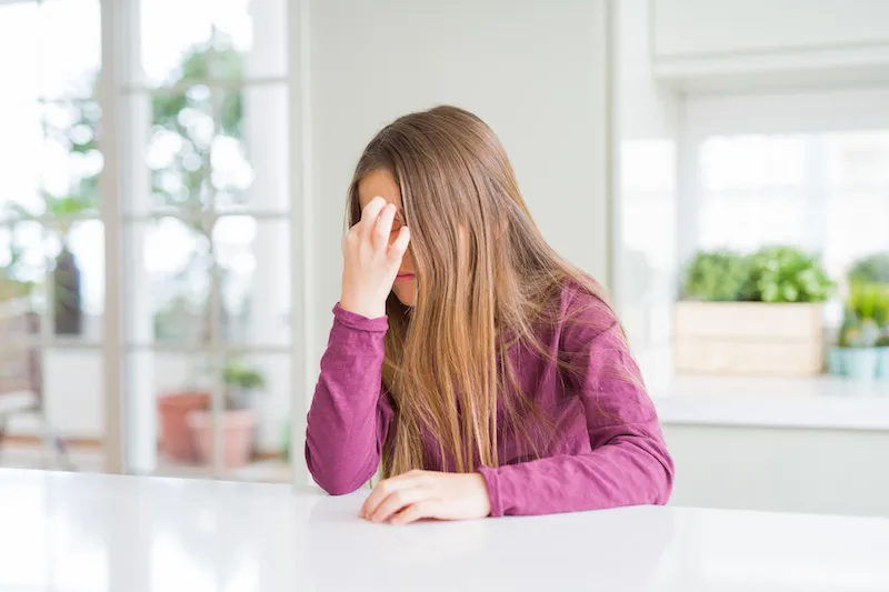 Seated girl in purple shirt holds hand against head, as if suffering a headache